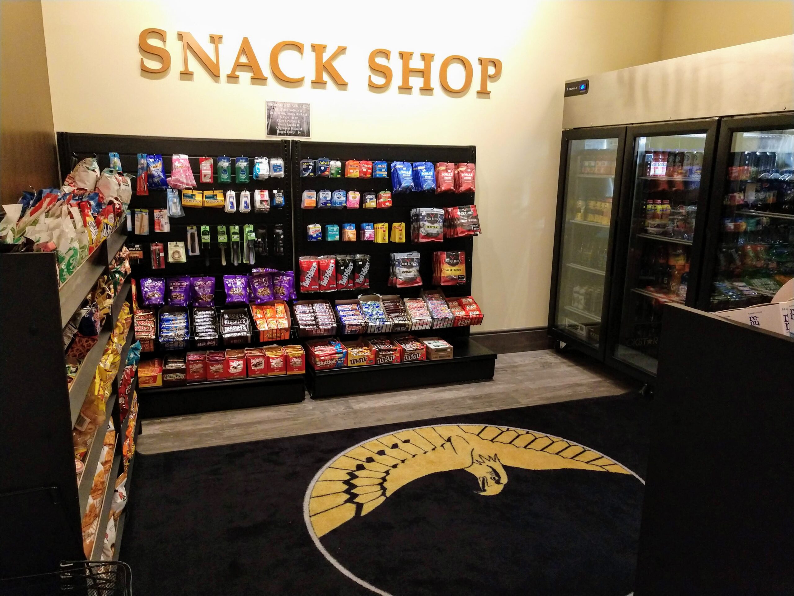 nterior of a snack shop featuring shelves filled with various snacks and drinks. The sign above reads "SNACK SHOP." A large rug with a logo is visible in the center of the floor, and glass refrigerators display cold beverages nearby.