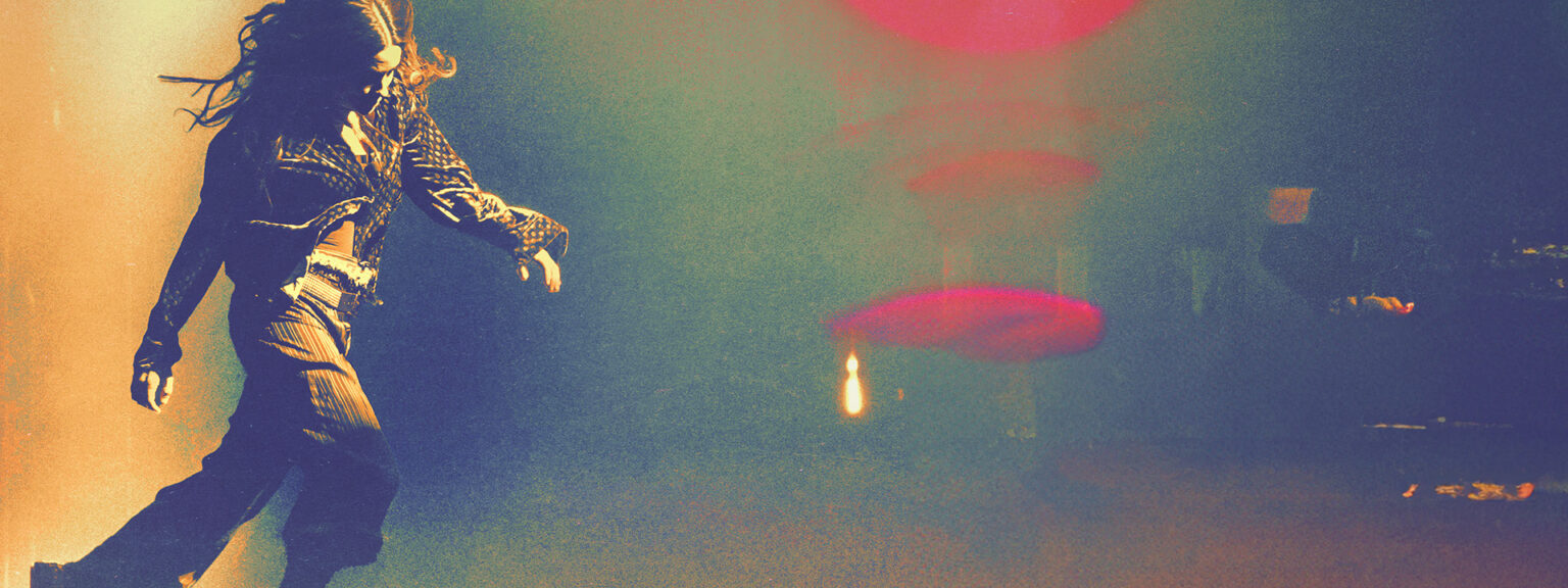 A performer in a dark venue, captured in motion, wearing a black leather jacket and loose pants. Colorful stage lights, including red and yellow hues, illuminate the background, enhancing the dynamic atmosphere of the scene.